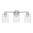 The Lyndsey - 60203, a three-light fixture from Craftmade, has brushed nickel finishes and clear textured glass cylindrical shades with slender bulbs. It features an oval backplate for a modern and sleek aesthetic.